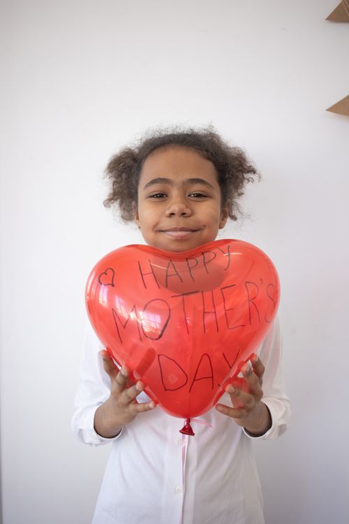 Free Smiling child holding a heart-shaped balloon with 'Happy Mother's Day' message against a white wall. Stock Photo