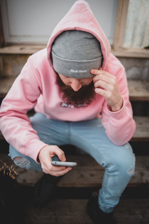 From above of male with beard in pink hoody and cup sitting on wooden stairs and browsing mobile phone