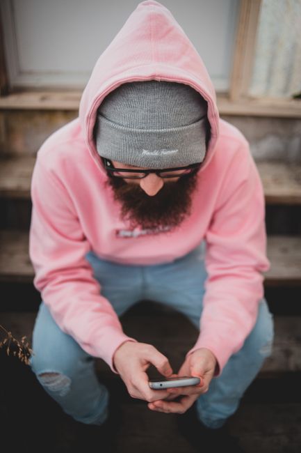 Bearded man wearing pink hoodie and gray beanie sits texting on smartphone, embodying casual urban style.