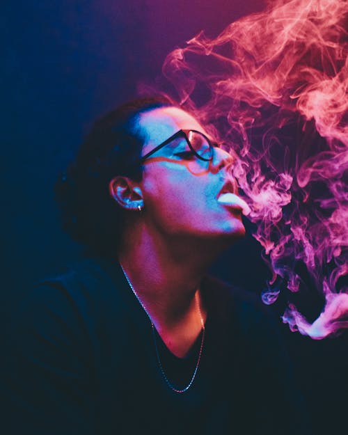 Male smoker in black outfit and eyeglasses exhaling smoke in air in room with bright neon lights