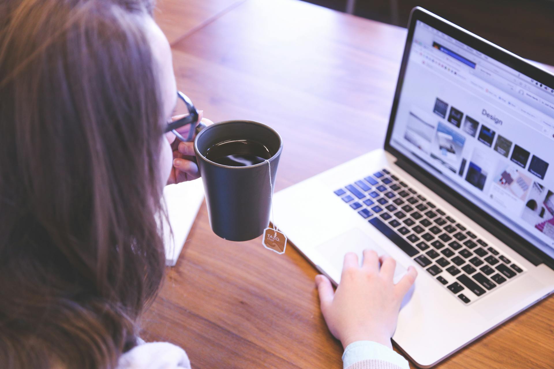 A woman enjoys coffee while browsing the web on a laptop at a home office desk.