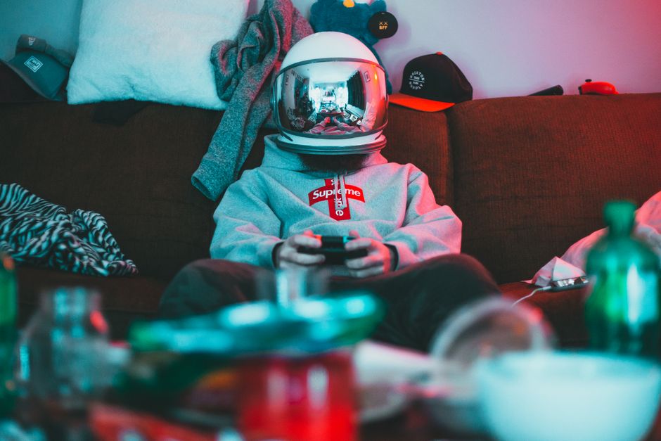 Concentrated teen in casual warm hoodie and trousers wearing virtual reality helmet sitting on couch surrounded by mess playing videogame