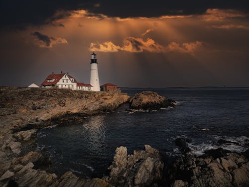 Picturesque view of building and lighthouse exteriors on rocky beach against ocean under cloudy sky at sunset