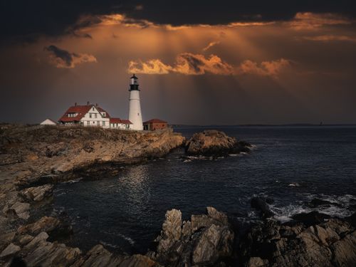 Free Picturesque view of building and lighthouse exteriors on rocky beach against ocean under cloudy sky at sunset Stock Photo