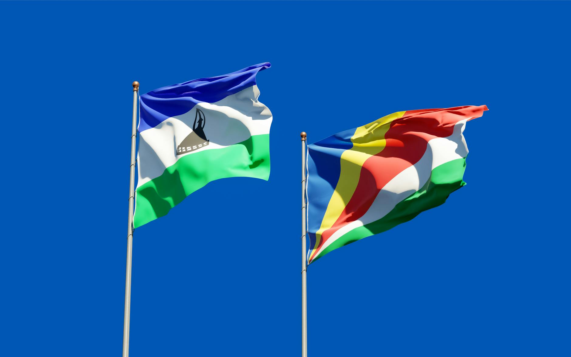 National flags of Lesotho and Seychelles waving on flagpoles against a clear blue sky.