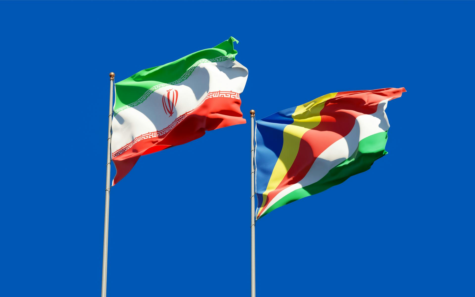 National flags of Iran and Seychelles waving on poles against a bright blue sky, symbolizing international relations.