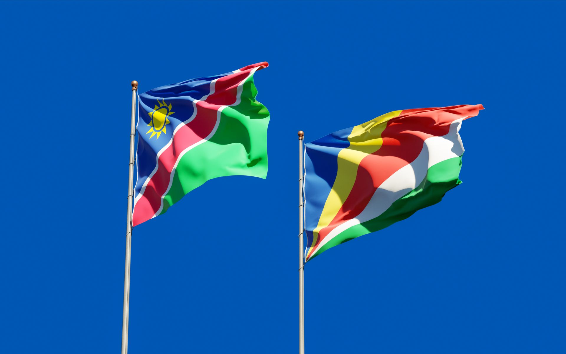 Colorful flags of Namibia and Seychelles waving on flagpoles against a clear blue sky.