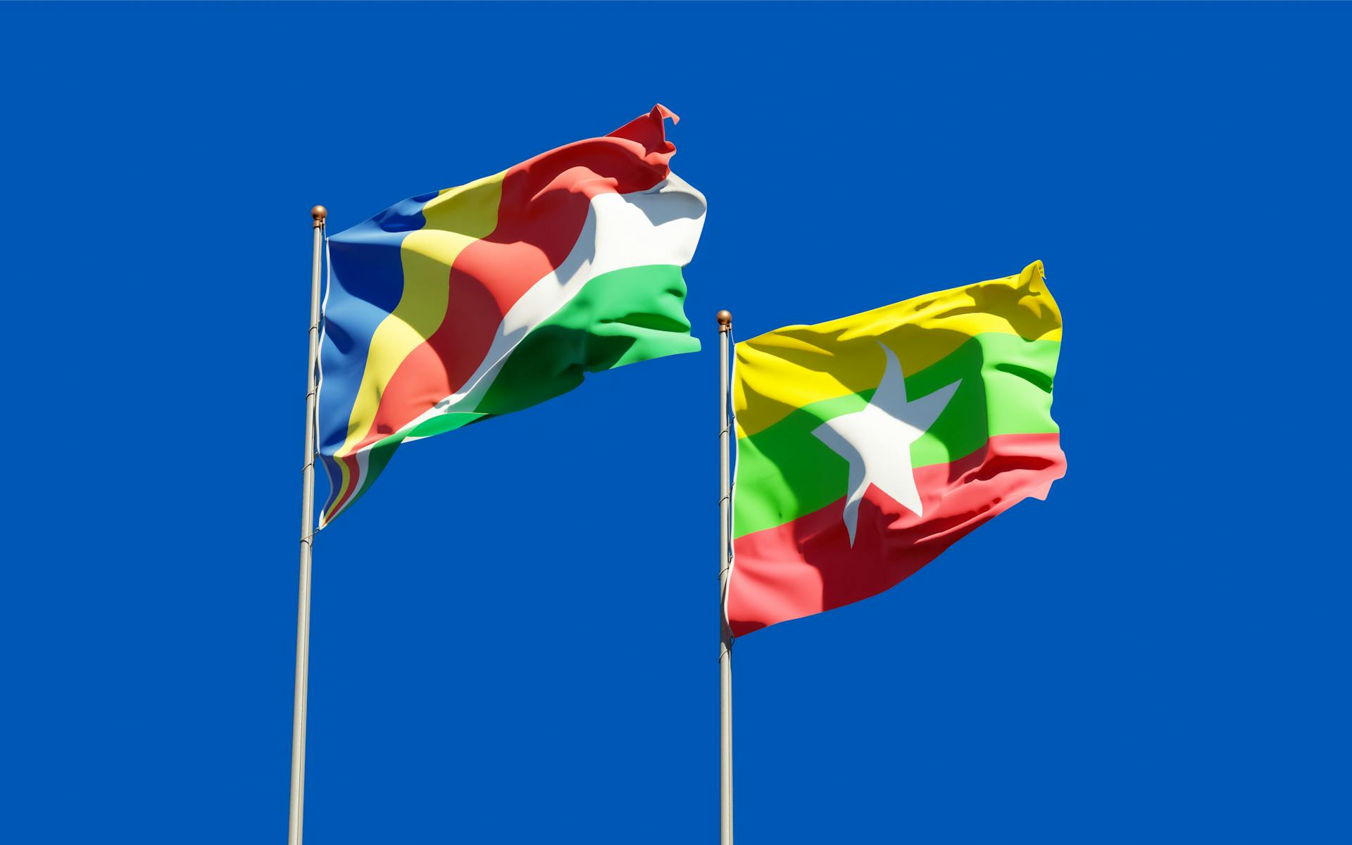 National flags of Seychelles and Myanmar waving on flagpoles against a clear blue sky.