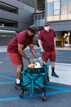 Two delivery men in masks arrange groceries in a green cart in a parking area