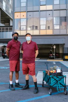Two delivery men in masks stand with a cart filled with groceries outdoors in a city.