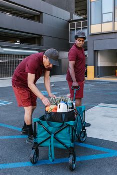 Two men in maroon uniforms deliver groceries in an urban parking lot.