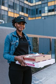 Female pizza delivery worker smiling while holding pizza boxes outside an urban building.
