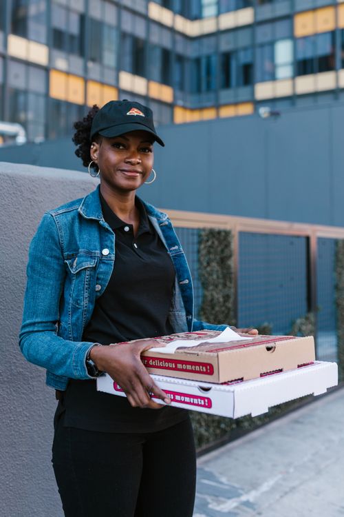 Free Female pizza delivery worker smiling while holding pizza boxes outside an urban building. Stock Photo