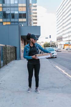 A delivery woman with pizza boxes stands on a city sidewalk, wearing a denim jacket and cap.