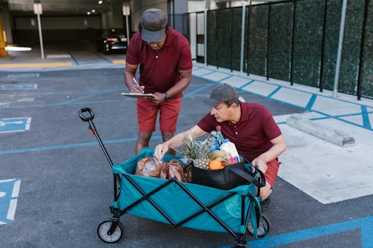 Two men in red uniforms delivering groceries with a push cart in a parking area.