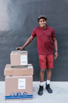 Smiling courier in maroon outfit with packages on sidewalk against dark wall.