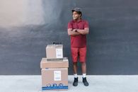 Man in Red Polo Shirt and Red Pants Standing Beside Brown Cardboard Boxes