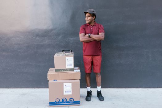 Delivery man in red attire posing outdoors with stacked cardboard boxes against a concrete wall.
