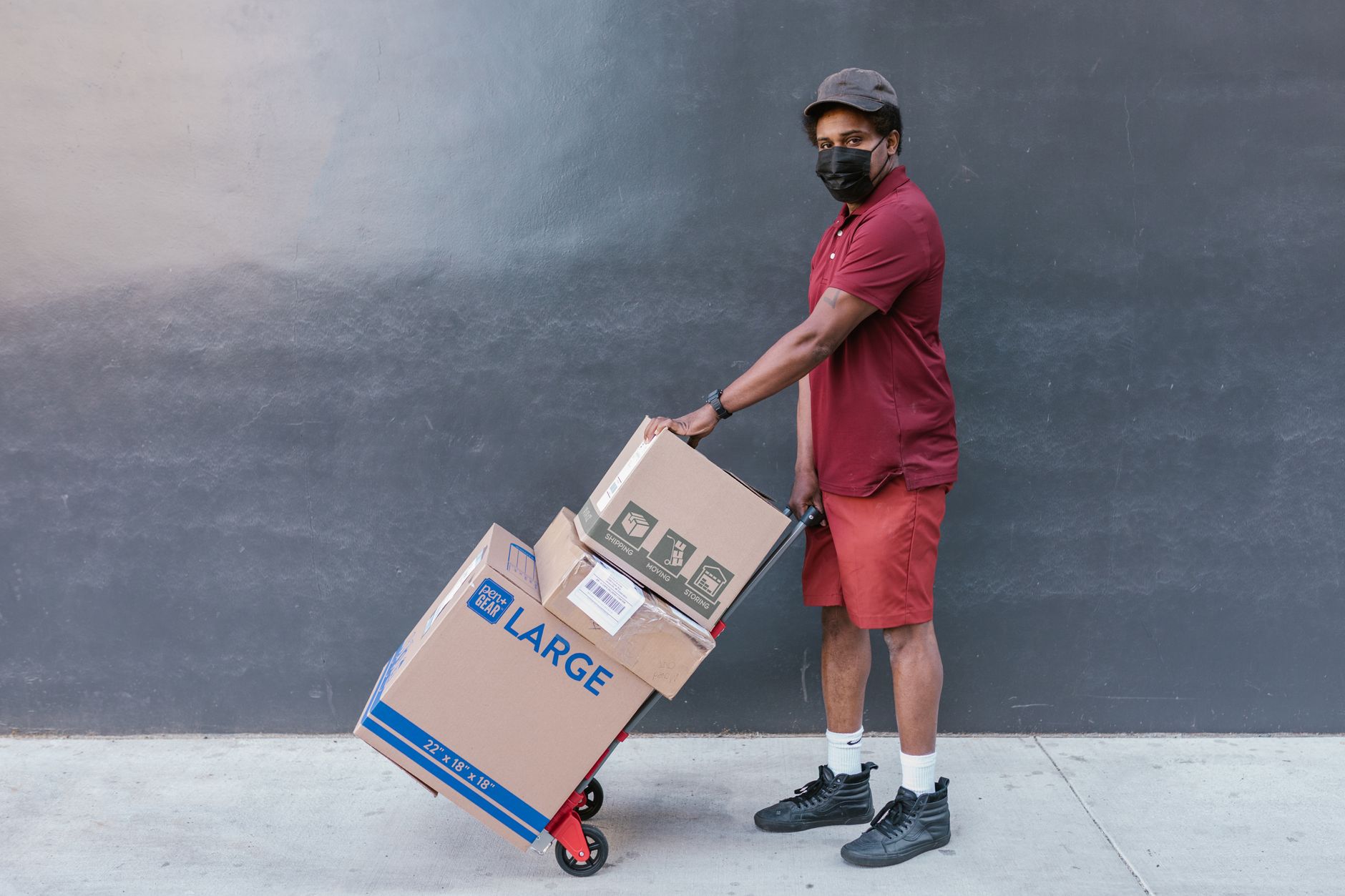Delivery worker wearing face mask transporting boxes on a cart outside. Safe and essential service during COVID-19 pandemic.