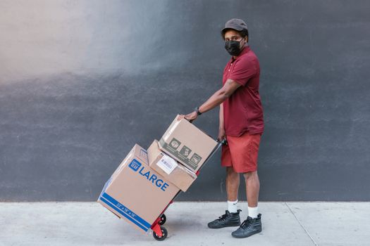 Delivery worker wearing face mask transporting boxes on a cart outside. Safe and essential service during COVID-19 pandemic.