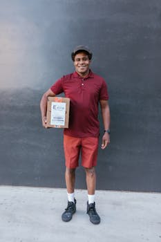 A smiling delivery man in a red polo shirt and shorts holds a package against a concrete wall.