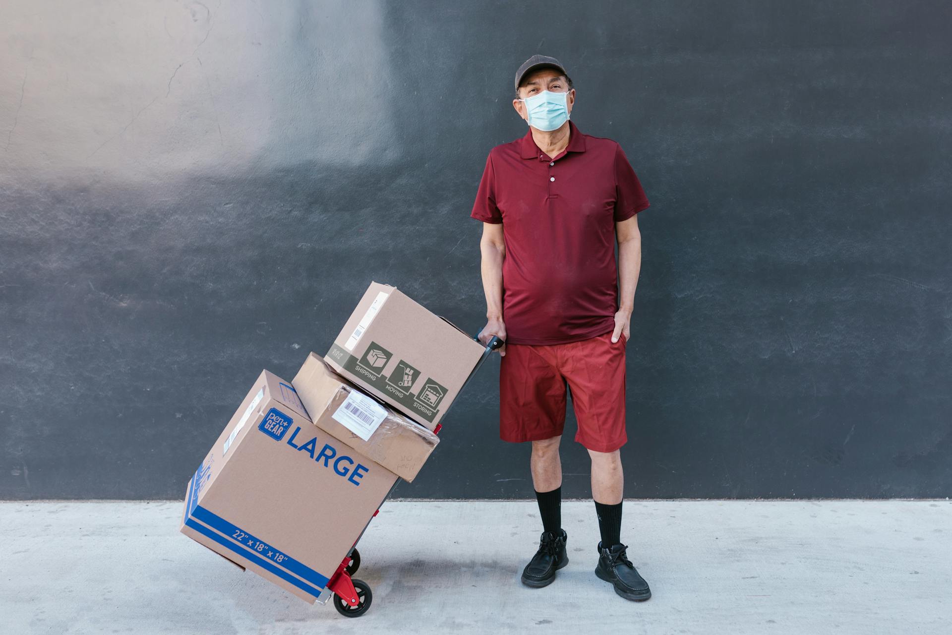 Courier in red uniform and face mask with boxes on trolley against a plain backdrop.