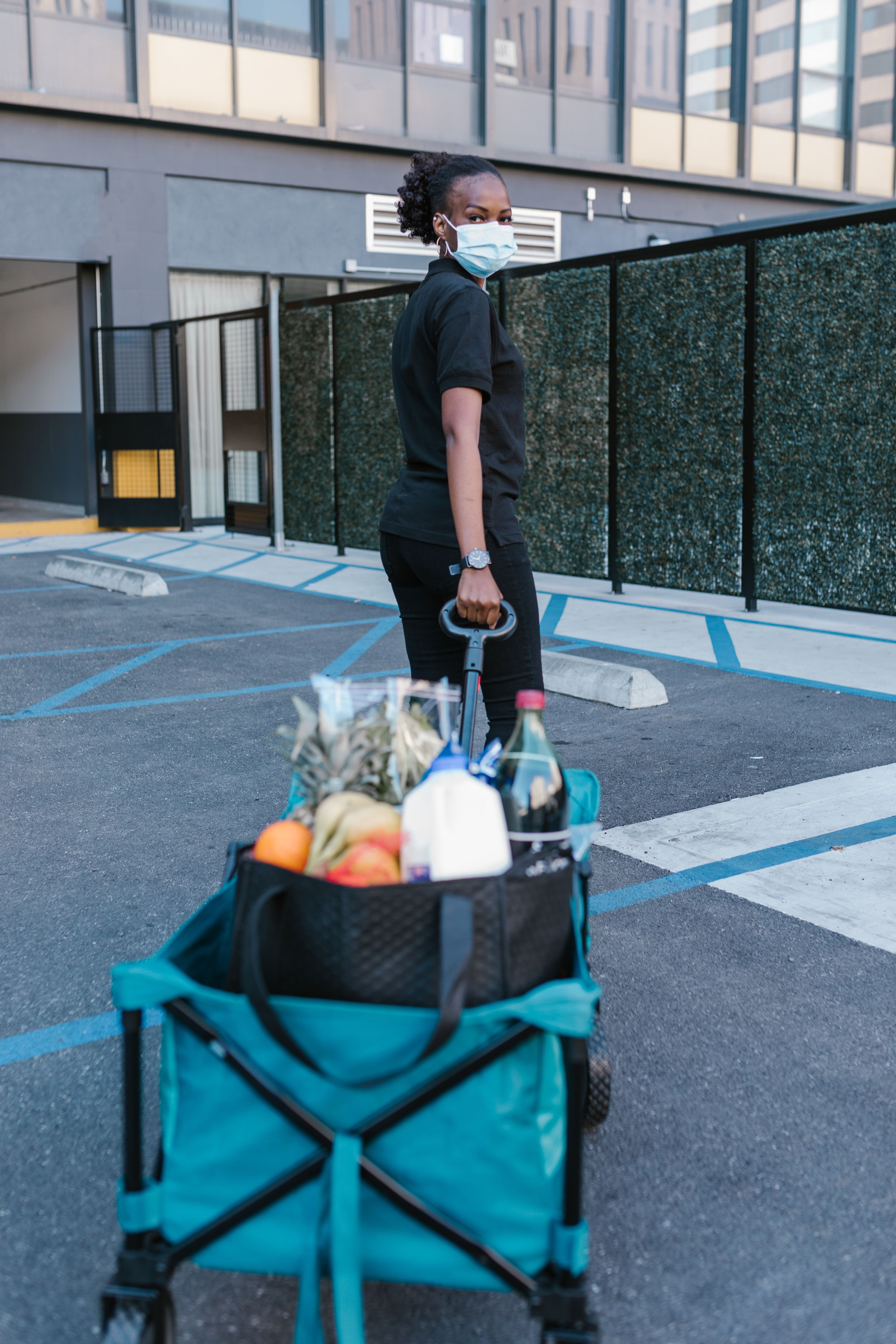 Focused deliverywoman in face mask pulling a cart with groceries through a parking lot.