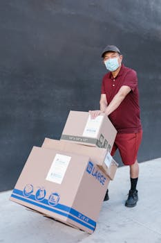 Delivery person with facemask pushing cart with boxes outdoors.