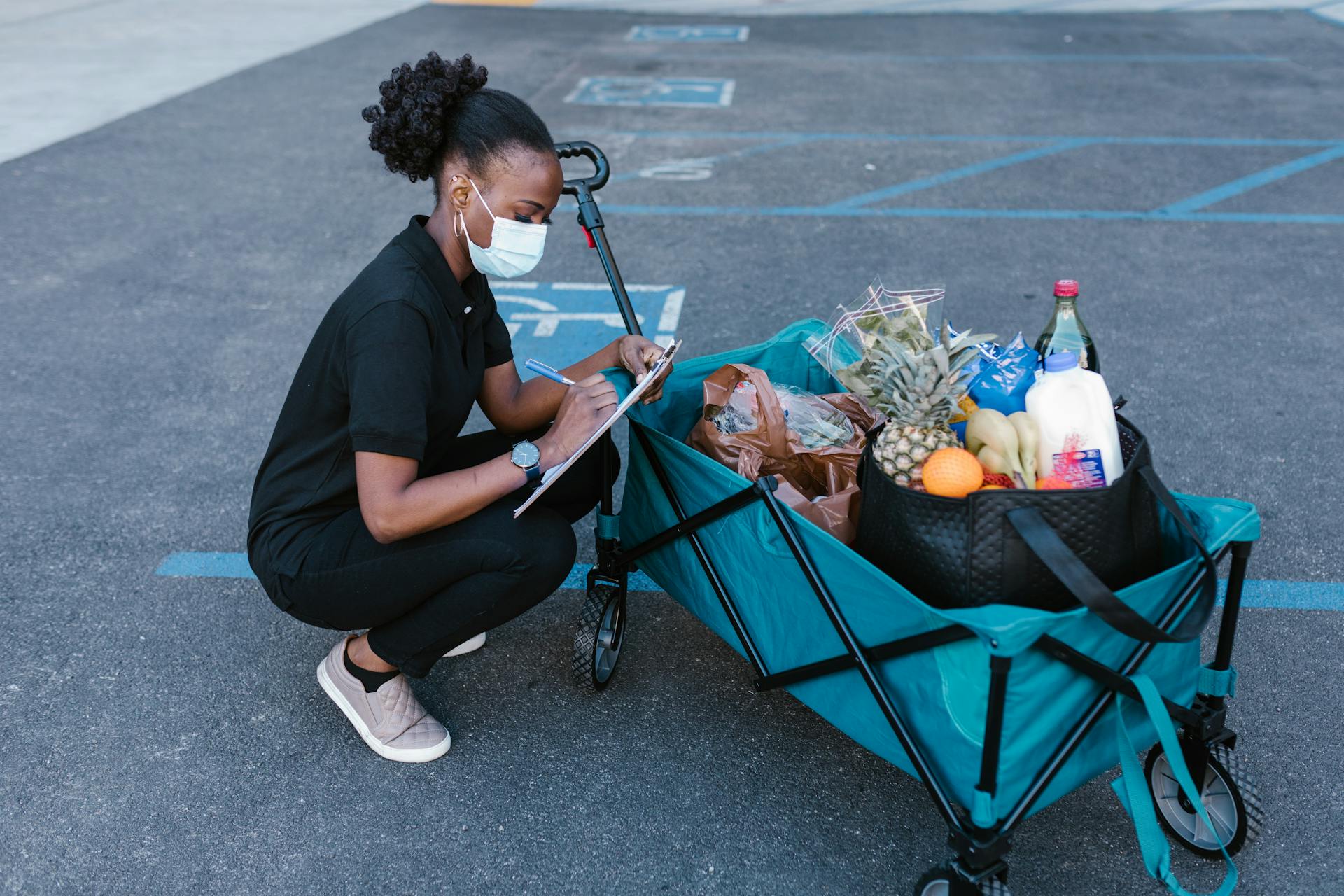 A woman in a face mask checking a clipboard while squatting next to a delivery cart full of groceries.