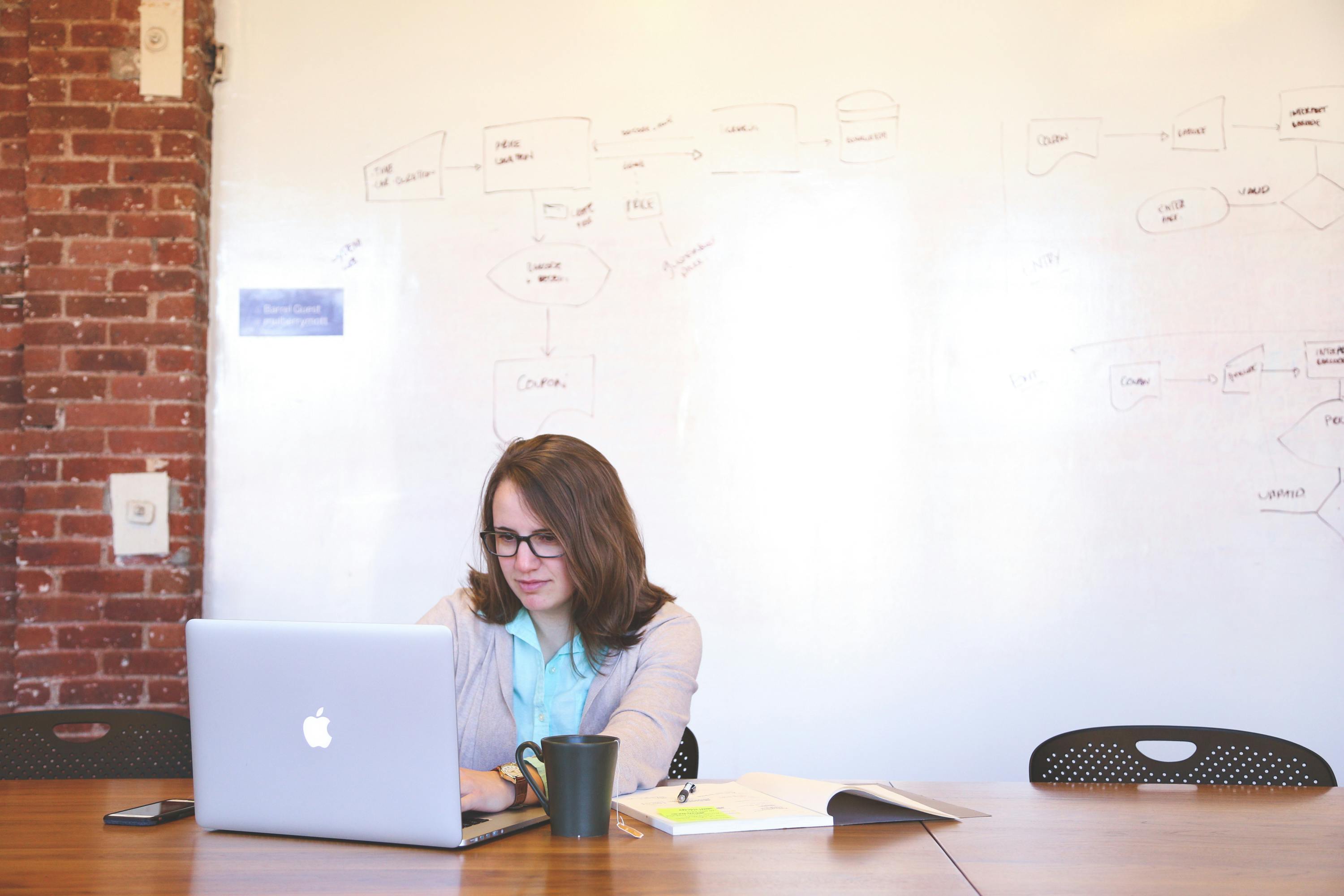 woman wearing black framed eyeglasses and teal button up shirt and beige blazer sitting at table near white macbook