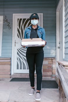 Pizza deliverywoman wearing mask stands at front porch with pizza boxes, portraying contactless delivery.