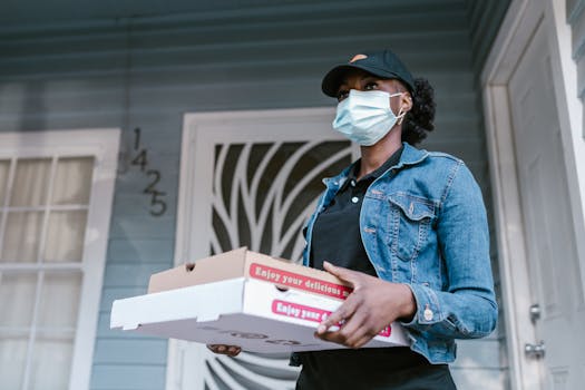 Delivery woman wearing a mask delivering pizza boxes at a front door. Outdoor scene.