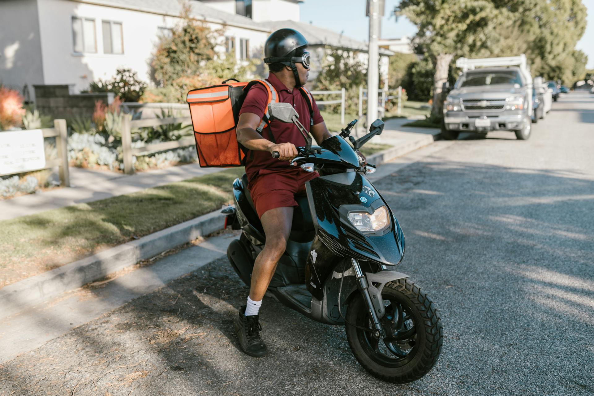 A delivery person on a scooter in a sunny neighborhood street, wearing a helmet and maroon uniform.