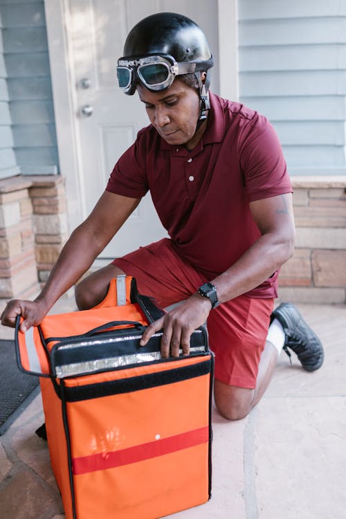 Homme En Polo Rouge Et Pantalon Gris Assis Sur Des Bagages Orange Et Noir