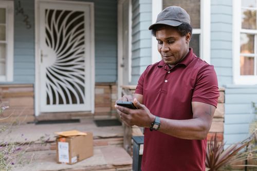 Free A courier in a maroon shirt checks delivery details on his phone at a residential porch with parcels. Stock Photo