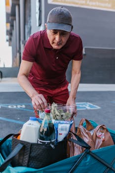A delivery man in a red polo shirt organizes groceries in bags outdoors, representing essential services.