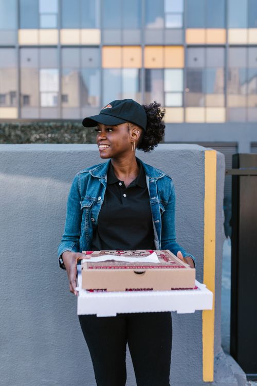 Free A cheerful delivery woman wearing a cap and denim jacket holds pizza boxes outdoors. Stock Photo