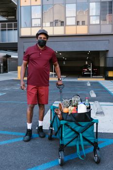 Delivery person with grocery cart in urban setting, wearing mask for pandemic safety.