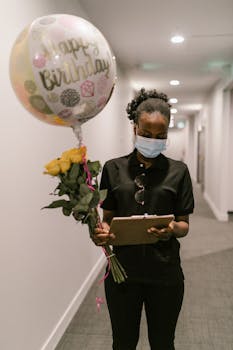 A delivery woman wearing a mask holds a birthday balloon and roses in a hallway.