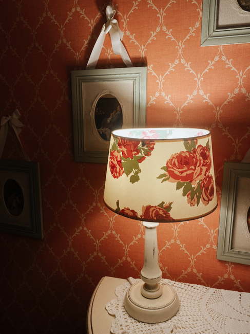 A warm vintage lamp with a floral shade illuminates an ornate orange patterned wall and framed pictures.