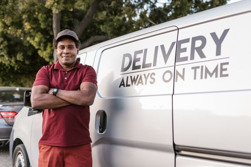 Free Delivery driver standing with arms crossed near a company van displaying a timely service slogan. Stock Photo