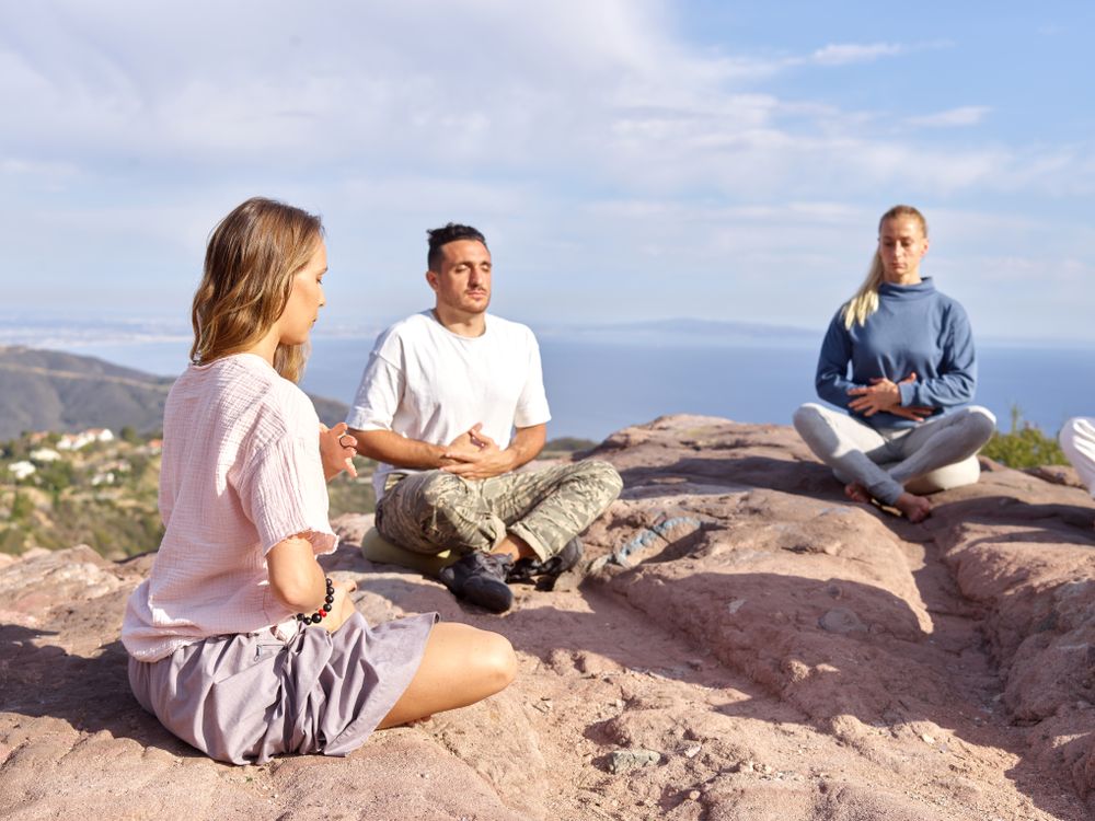 Three people practicing calming wellness exercises outdoors on a hillside