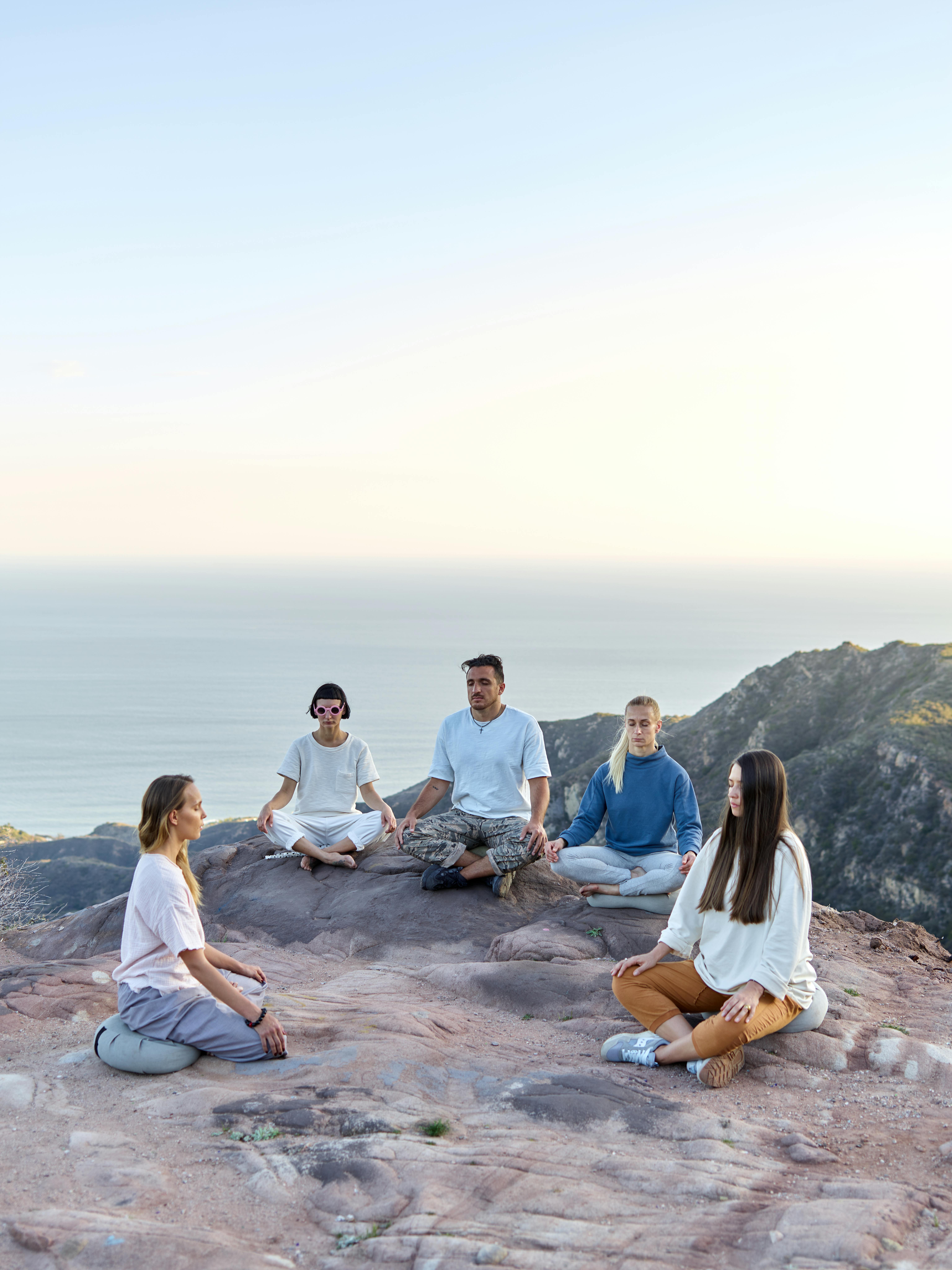 Diverse group meditating outdoors on a rocky cliff overlooking the ocean.
