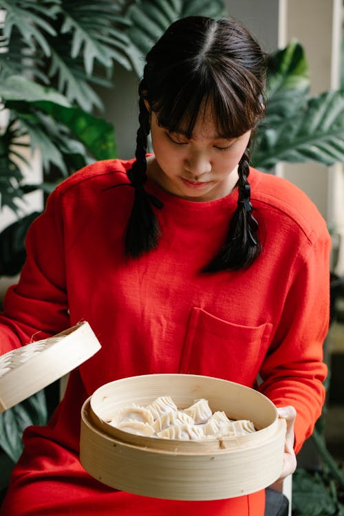 Young calm Asian female opening bamboo steamer and looking at set of delicious ready dumplings against plant
