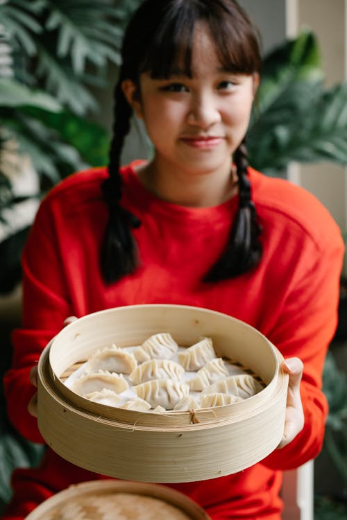 Crop ethnic teenager showing bamboo steamer with tasty dumplings while looking at camera in house