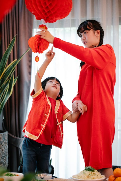 Ethnic female teenager in red dress holding sibling by wrist while decorating home with lanterns during New Year holiday