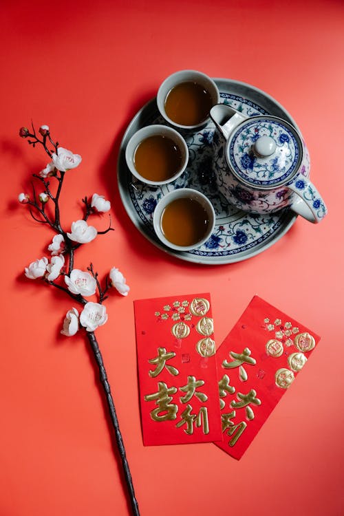 Traditional Chinese tea set with cups and teapot on a tray, accompanied by red envelopes and a blossoming branch on a red background