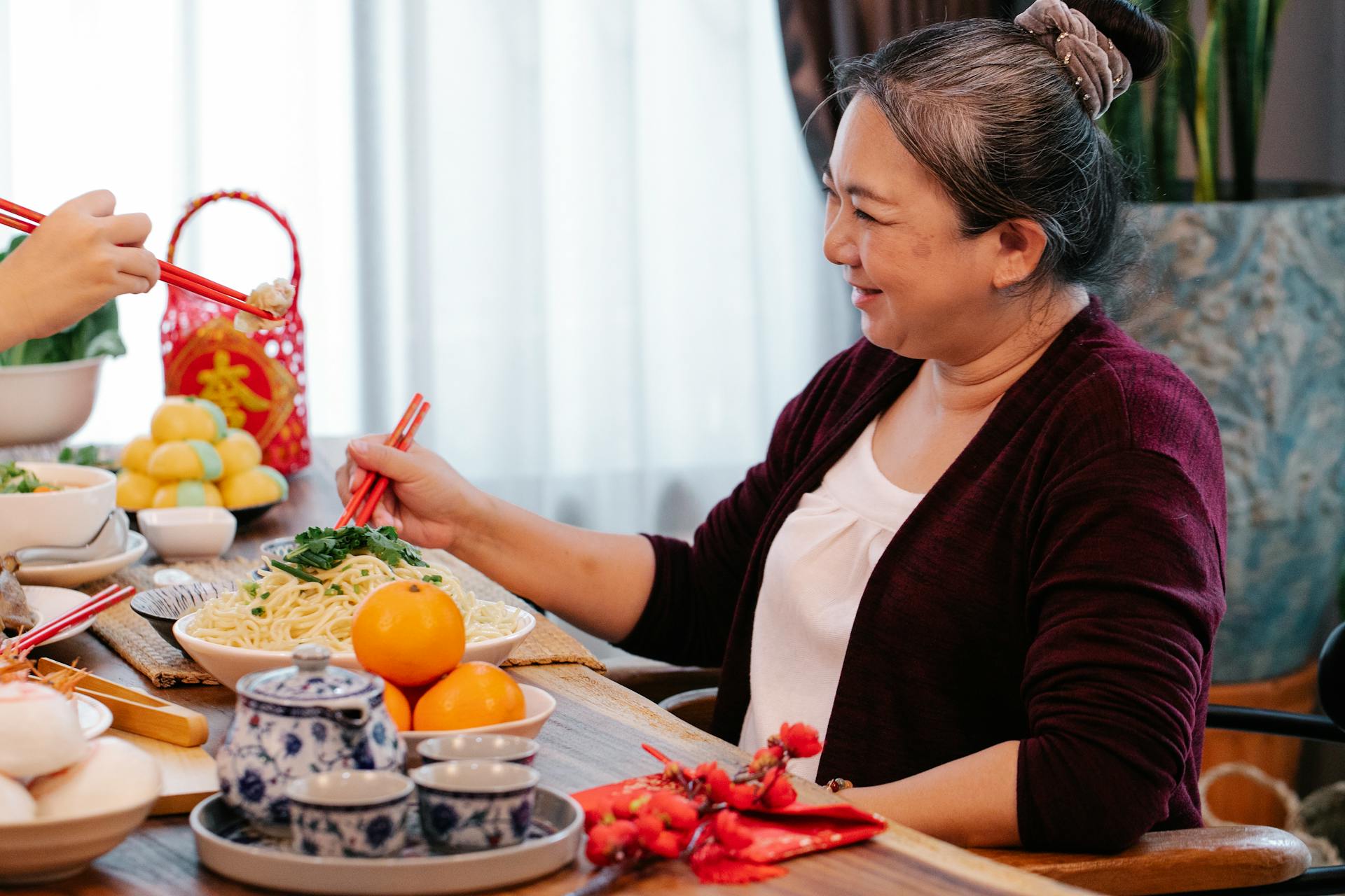 Cheerful mature ethnic female talking to crop unrecognizable relative at table with delicious noodles during New Year holiday at home