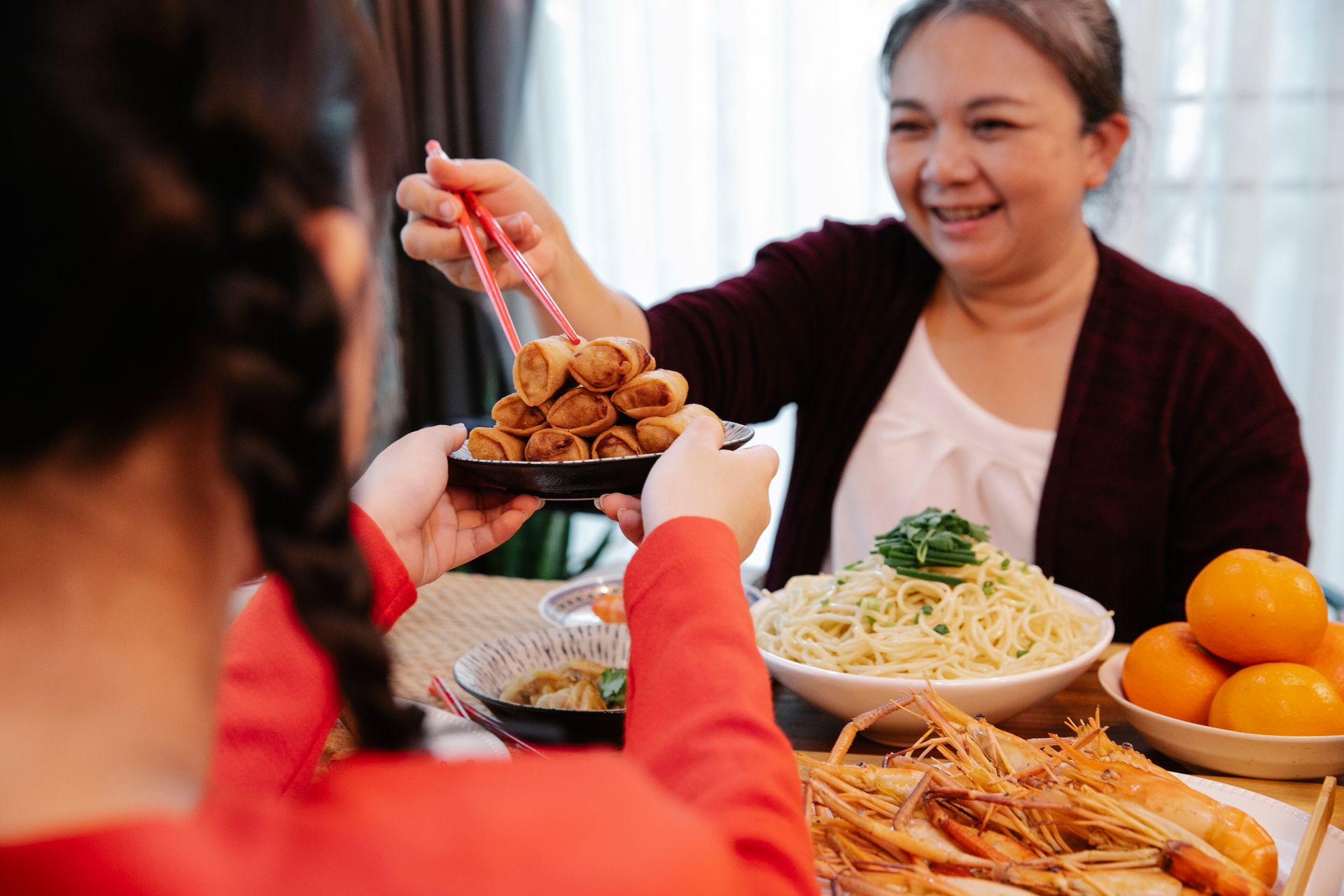 Crop unrecognizable female teenager serving delicious oriental rolls to smiling ethnic grandma with food sticks in house