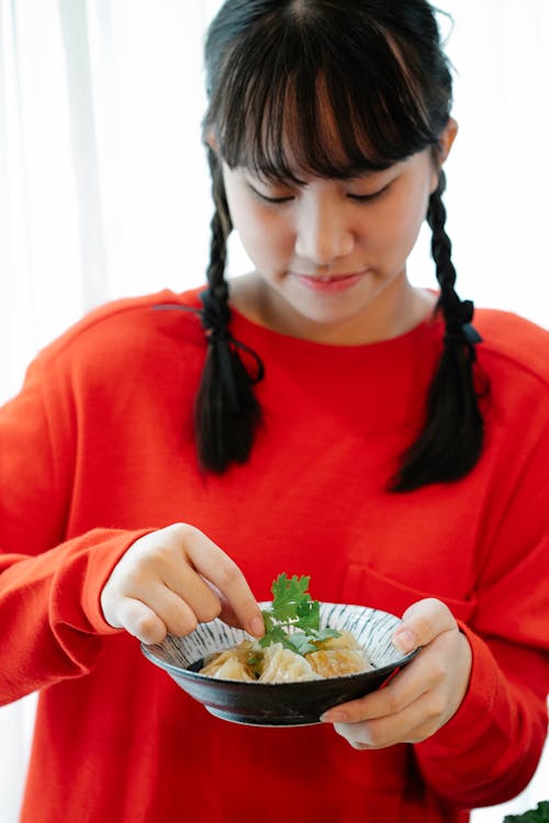 Crop ethnic female teenager in red wear with bowl of tasty Chinese dumplings with fresh parsley leaves on top